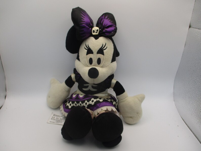 Puede incluir: Mu&ntilde;eca de peluche de Minnie Mouse con disfraz de Halloween. La mu&ntilde;eca tiene una combinaci&oacute;n de colores en blanco y negro con un lazo y una falda morados. El lazo presenta una calavera. La mu&ntilde;eca tiene zapatos y guantes negros.