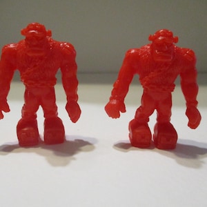 Puede incluir: Dos figuras rojas de monstruos musculosos. Cada figura tiene una cabeza grande, hombros anchos y piernas gruesas. Las figuras están de pie con los brazos a los lados. Las figuras son idénticas y proyectan una sombra sobre la superficie blanca.