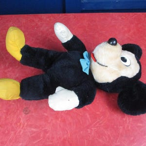 Op de afbeelding: Een vintage Mickey Mouse pluche speelgoed, liggend op zijn rug. Het pluche heeft een zwarte body, witte handschoenen en voeten en gele schoenen. Het heeft een cr&egrave;mekleurig gezicht, zwarte oren en een blauwe vlinderdas. Het speelgoed ligt op een rode ondergrond.