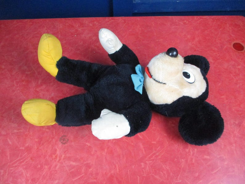 Op de afbeelding: Een vintage Mickey Mouse pluche speelgoed, liggend op zijn rug. Het pluche heeft een zwarte body, witte handschoenen en voeten en gele schoenen. Het heeft een cr&egrave;mekleurig gezicht, zwarte oren en een blauwe vlinderdas. Het speelgoed ligt op een rode ondergrond.