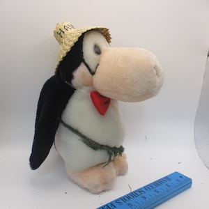 May include: A plush penguin toy with a straw hat that says "HAWAI" and a red bow tie. The penguin has black wings and a green grass skirt. The penguin is standing on a white surface.