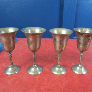 Pode incluir: Quatro cálices vintage prateados com uma taça em forma de tulipa, haste fina e base redonda. Os cálices estão dispostos em fila sobre uma superfície vermelha, com um fundo azul. Os cálices mostram sinais de idade e desgaste.