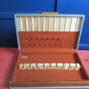 May include: Open silverware storage case with a light blue frame, orange interior, and cream-colored satin lining. The case has slots for silverware and a small orange handle. The case is open, revealing the interior.