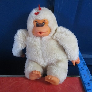 May include: A plush white gorilla toy with orange hands and feet. The gorilla has a brown face, a red heart on its head, and is sitting on a red surface. A blue ruler is next to the toy.