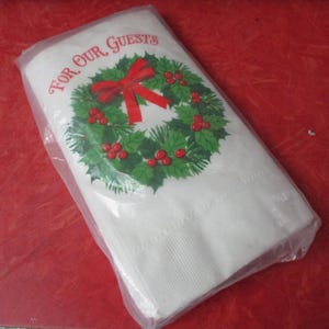 May include: A package of white paper napkins with a Christmas wreath design. The wreath is green with red berries and a red bow. The text "FOR OUR GUESTS" is printed above the wreath in red. The napkins are in a clear plastic wrapper.