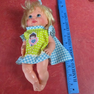 May include: A vintage doll with blonde hair and blue eyes, wearing a green and white polka-dot dress with a blue and white gingham trim. The doll has a blue and white gingham cape. A blue ruler is next to the doll.