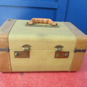 Puede incluir: Una maleta vintage con cuerpo amarillo claro y ribetes de cuero marrón. La maleta tiene un asa marrón, herrajes de latón y dos cierres. Las esquinas están reforzadas con cuero marrón y los bordes están cosidos con hilo blanco.