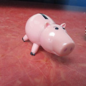 May include: A pink piggy bank with black eyes and feet. The piggy bank has a slot on its back for inserting coins. A toy figure of a cowgirl is in the background.