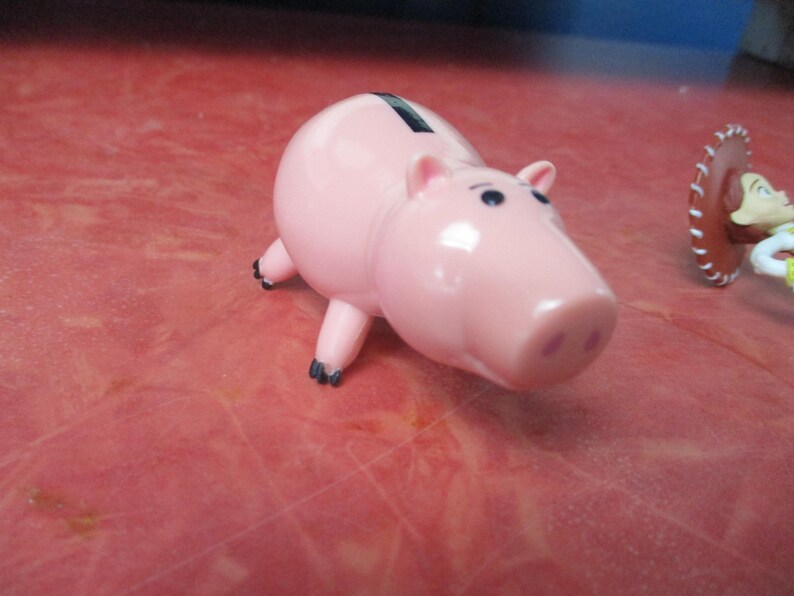 May include: A pink piggy bank with black eyes and feet. The piggy bank has a slot on its back for inserting coins. A toy figure of a cowgirl is in the background.