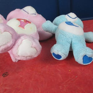 May include: Two plush teddy bear toys in the image. One is light pink with a heart on its foot and a design on its belly. The other is light blue with a heart on its foot and belly. Both have white accents.