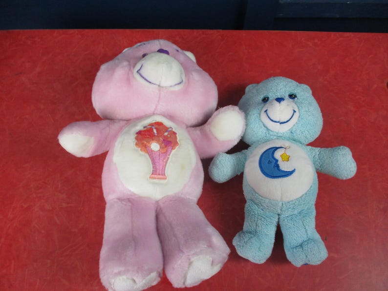 May include: Two plush Care Bears. One is light pink with a flower design on its belly. The other is light blue with a moon and star design. Both have smiling faces and are on a red surface.