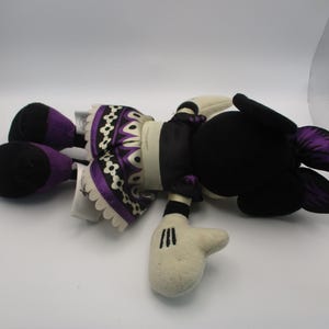 Puede incluir: Una mu&ntilde;eca de peluche con cabeza y orejas negras, cara y manos blancas. La mu&ntilde;eca tiene un cuerpo negro con una falda estampada en morado y blanco y detalles en morado en los pies y las orejas. La mu&ntilde;eca est&aacute; tumbada boca arriba.