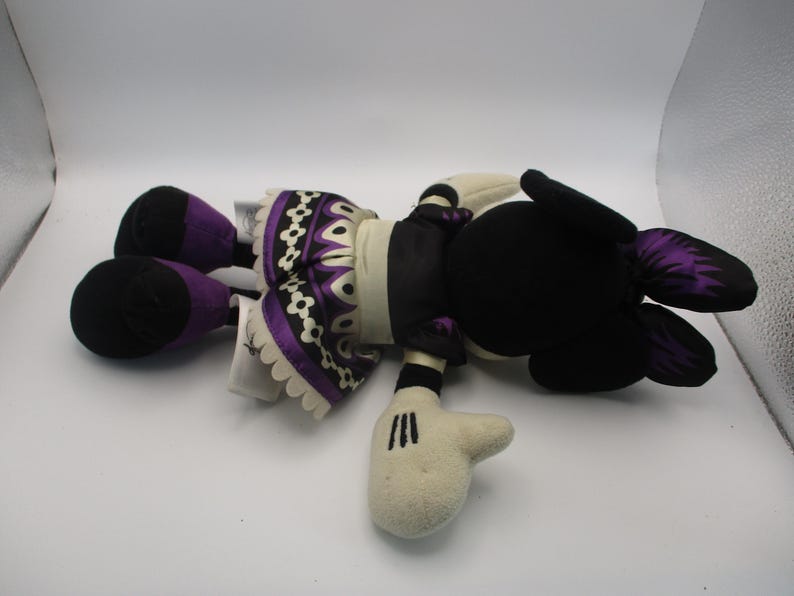 Puede incluir: Una mu&ntilde;eca de peluche con cabeza y orejas negras, cara y manos blancas. La mu&ntilde;eca tiene un cuerpo negro con una falda estampada en morado y blanco y detalles en morado en los pies y las orejas. La mu&ntilde;eca est&aacute; tumbada boca arriba.