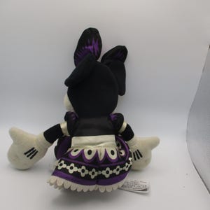 Puede incluir: Mu&ntilde;eca de peluche de un personaje, vista desde atr&aacute;s. La mu&ntilde;eca tiene orejas negras con detalles morados, una cabeza negra y una cara blanca. Viste un vestido negro con un delantal blanco y una falda estampada morada y blanca. La mu&ntilde;eca tiene guantes blancos con rayas negras.