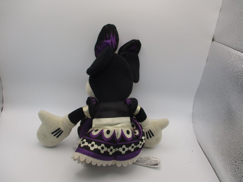 Puede incluir: Mu&ntilde;eca de peluche de un personaje, vista desde atr&aacute;s. La mu&ntilde;eca tiene orejas negras con detalles morados, una cabeza negra y una cara blanca. Viste un vestido negro con un delantal blanco y una falda estampada morada y blanca. La mu&ntilde;eca tiene guantes blancos con rayas negras.