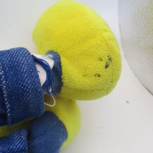 May include: A plush toy with a yellow head and blue denim clothing. The toy's head is a bright yellow color, and the denim clothing is a deep blue. The toy appears to be well-loved, with some wear and tear visible.