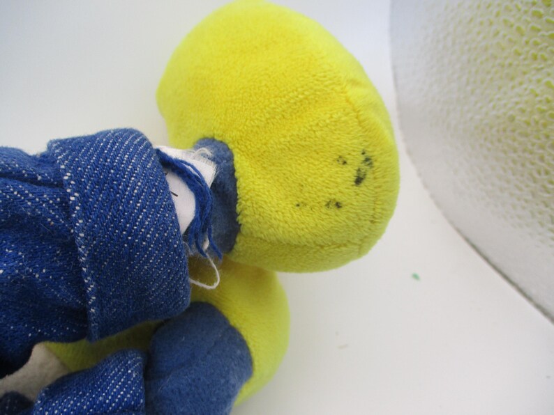 May include: A plush toy with a yellow head and blue denim clothing. The toy's head is a bright yellow color, and the denim clothing is a deep blue. The toy appears to be well-loved, with some wear and tear visible.