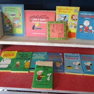 May include: A collection of vintage Peanuts books, including titles like "Good Ol' Snoopy" and "You're the Greatest, Charlie Brown," displayed on a white shelf and a red surface. The books feature colorful covers with illustrations of Snoopy, Charlie Brown, and other characters.