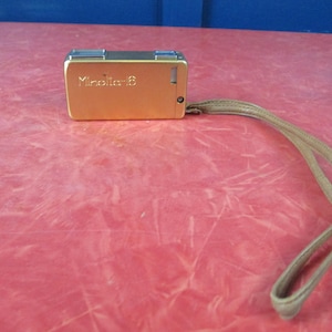 May include: A vintage gold-colored Minolta-16 camera with a matching wrist strap. The camera is rectangular with a small lens and a button on the side. The wrist strap is a light tan color.