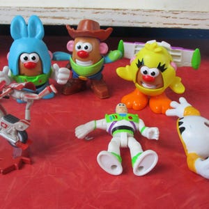 May include: A selection of colourful Toy Story character figurines, including Mr. Potato Head in various costumes, Buzz Lightyear, and a yellow chick. A toy motorcycle is also present. The toys are arranged on a red surface.