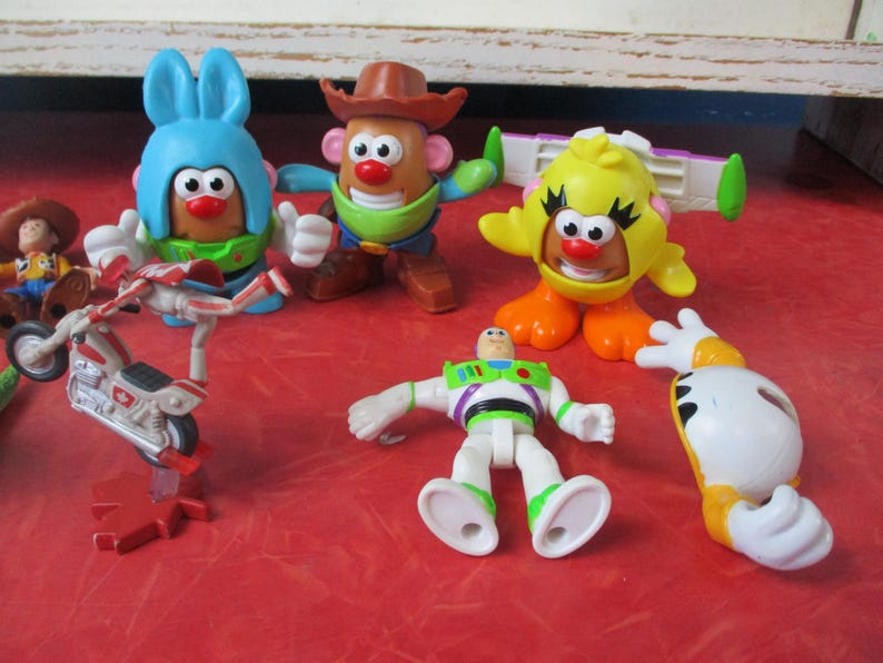 May include: A selection of colourful Toy Story character figurines, including Mr. Potato Head in various costumes, Buzz Lightyear, and a yellow chick. A toy motorcycle is also present. The toys are arranged on a red surface.