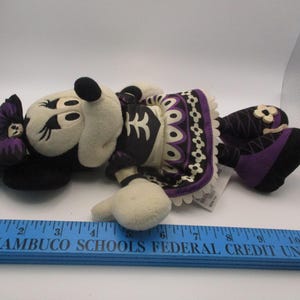 Puede incluir: Mu&ntilde;eca de peluche de Minnie Mouse con tem&aacute;tica del D&iacute;a de Muertos. La mu&ntilde;eca presenta una cara y manos color crema, un lazo negro y un vestido negro y morado con adornos decorativos. La mu&ntilde;eca mide aproximadamente 28 cm de largo.
