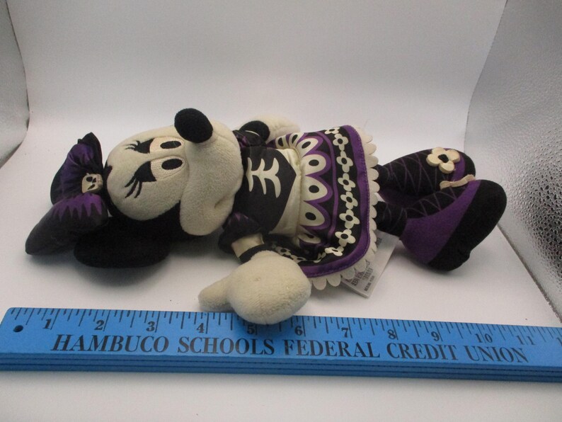 Puede incluir: Mu&ntilde;eca de peluche de Minnie Mouse con tem&aacute;tica del D&iacute;a de Muertos. La mu&ntilde;eca presenta una cara y manos color crema, un lazo negro y un vestido negro y morado con adornos decorativos. La mu&ntilde;eca mide aproximadamente 28 cm de largo.