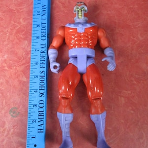 May include: A red and purple action figure of a superhero, standing upright. The figure has a red torso and legs, with purple accents on the arms, waist, and boots. A blue ruler is next to the figure, with the text "HAMBUCO SCHOOLS FEDERAL CREDIT UNION".