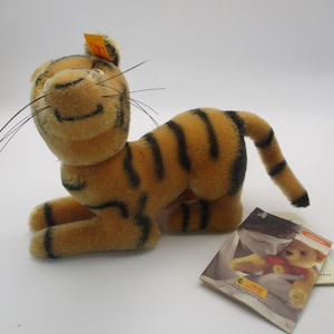 May include: A plush toy tiger in a seated position, featuring a tan body with black stripes. The tiger has long whiskers and a smiling face. A tag is attached to the ear. A small card with a picture of a teddy bear is next to the tiger.