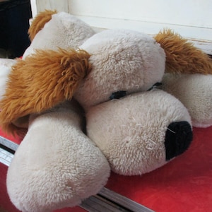 Op de afbeelding: Een crème en bruin pluche hondenspeelgoed. De hond heeft bruine oren, een zwarte neus en een rode hart op een poot. Het speelgoed ligt op een rode ondergrond.