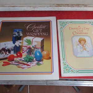 May include: Two books on a red surface. One is a spiral-bound book titled "Creative Gift Wrapping" with images of wrapped gifts. The other is titled "A Devotion a Day Until Christmas" with an illustration of an angel.