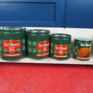 May include: Four vintage Del Monte brand food cans of varying sizes, arranged on a white shelf. The cans are dark green with red and white labels. The labels indicate the contents: tomatoes, whole string beans, yellow cling peaches, and mixed fruit.