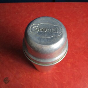 Könnte beinhalten: Ein Vintage-Cocamalt-Behälter aus Metall mit silbernem Finish. Das Wort "Cocamalt" ist auf dem Deckel geprägt. Der Behälter ist zylindrisch mit abgerundeter Oberseite und steht auf einer roten Oberfläche.