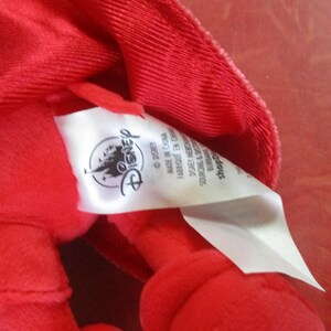 May include: Close-up of a bright red plush toy with a tag featuring the Disney logo. The tag reads "Made in China" and other text. The plush toy is made of a soft, textured fabric.