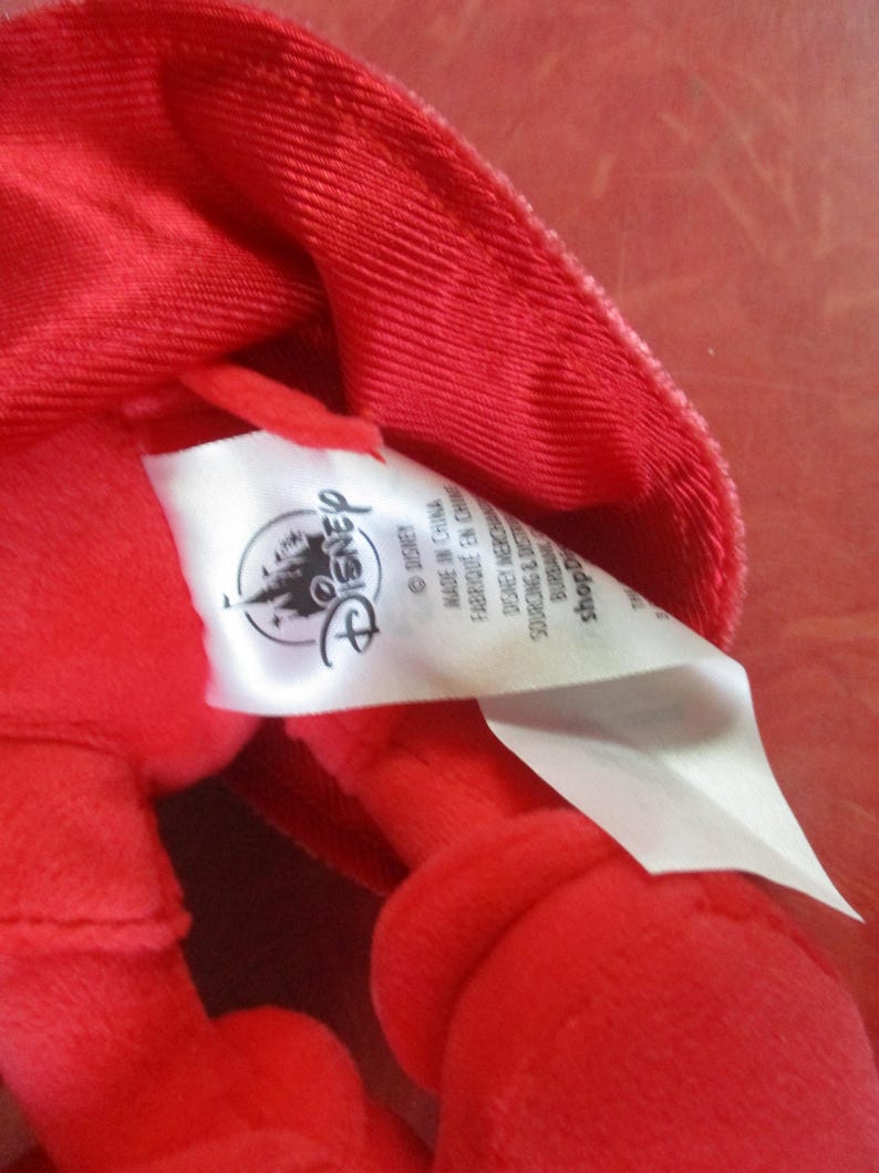 May include: Close-up of a bright red plush toy with a tag featuring the Disney logo. The tag reads "Made in China" and other text. The plush toy is made of a soft, textured fabric.