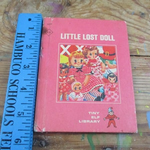 May include: A vintage pink children's book titled "Little Lost Doll" with illustrations of dolls on the cover. A blue ruler with white markings and the text "Hambuco Schools Fel" is on the left side of the book.