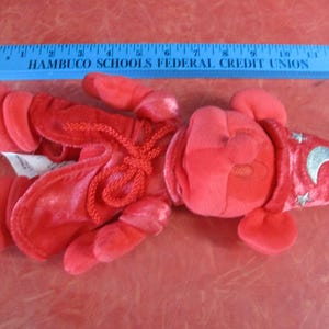 May include: A red plush toy in the shape of a lobster, with a wizard hat featuring a silver crescent moon and stars. The toy is lying on a red surface. A blue ruler is above the toy.