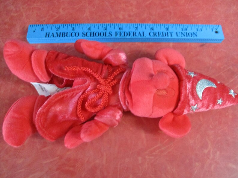 May include: A red plush toy in the shape of a lobster, with a wizard hat featuring a silver crescent moon and stars. The toy is lying on a red surface. A blue ruler is above the toy.