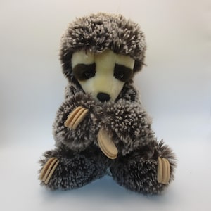 May include: A plush hedgehog toy with a light brown face, dark brown eyes, and a fluffy, textured body in shades of brown and gray. The toy has small, wooden-like feet and is sitting upright.