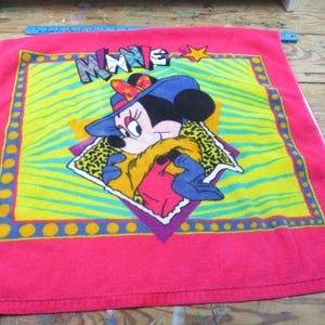 May include: A pink towel featuring Minnie Mouse in a blue hat and leopard print scarf. The design includes the word "MINNIE" in colorful letters, set against a yellow and green zebra-striped background, with a star.