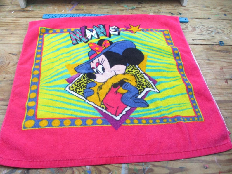 May include: A pink towel featuring Minnie Mouse in a blue hat and leopard print scarf. The design includes the word "MINNIE" in colorful letters, set against a yellow and green zebra-striped background, with a star.