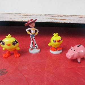 May include: A collection of Toy Story character figurines, including Jessie in a cowgirl outfit, two yellow chick figures, and a pink piggy bank. The figures are set on a red surface.