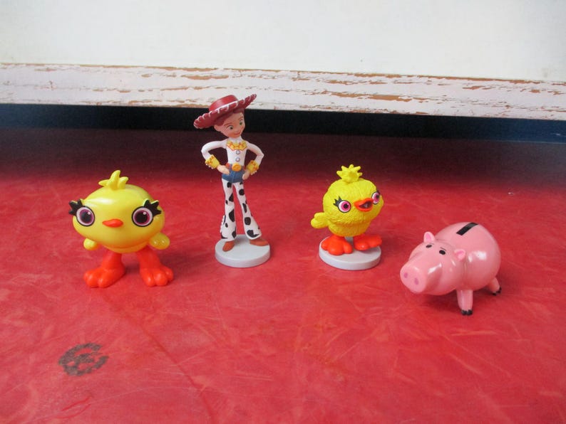 May include: A collection of Toy Story character figurines, including Jessie in a cowgirl outfit, two yellow chick figures, and a pink piggy bank. The figures are set on a red surface.