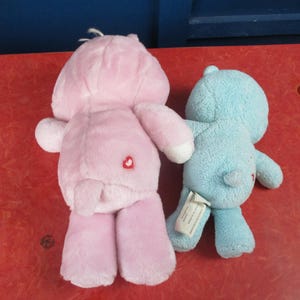 May include: Two plush toys: a pink bear with a heart on its rear and a blue bear. The pink bear has a heart on its rear. Both toys are on a red surface. The toys are soft and cuddly.