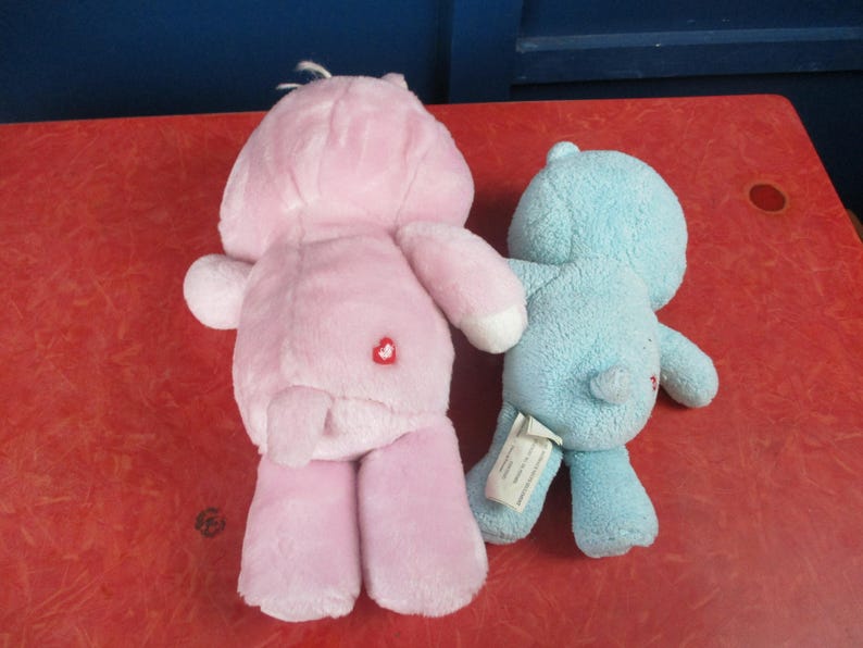 May include: Two plush toys: a pink bear with a heart on its rear and a blue bear. The pink bear has a heart on its rear. Both toys are on a red surface. The toys are soft and cuddly.