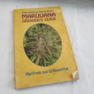 Può includere: Un libro giallo intitolato "Marijuana Grower's Guide" di Mel Frank e Ed Rosenthal. La copertina presenta una pianta di cannabis verde e viola all'interno di un ovale. Il libro è leggermente usurato.