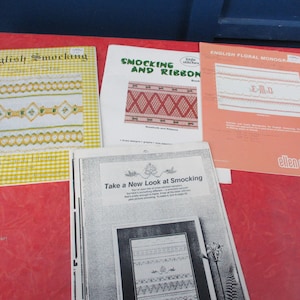 May include: A collection of vintage sewing and embroidery books. Titles include "English Smocking", "Smocking and Ribbon", and "English Floral Monogram". The books are on a red surface.