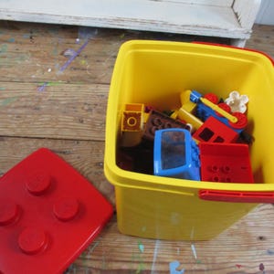 May include: A square yellow plastic storage bucket with a red handle and a separate red lid featuring four raised circular studs. The bucket is filled with various colorful plastic building blocks, including red, blue, and brown pieces, and a white flower.