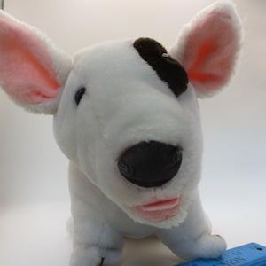 May include: A plush white dog with pink-lined ears and a black spot on its head. The dog has a black nose and a pink mouth. A blue ruler is in the foreground.