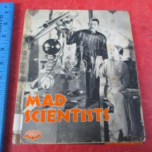 May include: Vintage book cover titled "Mad Scientists" featuring a black and white image of a man and woman with a complex machine. The title is in orange letters. The book is on a red surface.